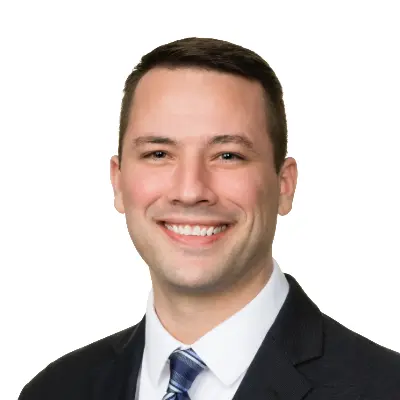 A man in a dark suit and striped tie smiles at the camera against a plain white background.
