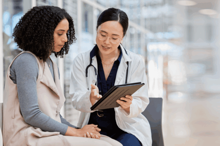 A doctor in a white coat uses predictive analytics to show information on a digital tablet to a seated woman, having a consultation in a modern, well-lit setting—a sign of how healthcare companies empower care with technology.