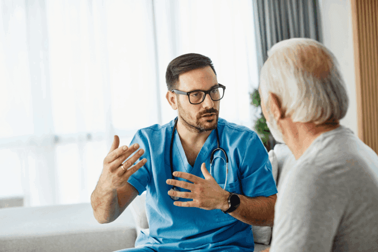A healthcare professional in blue scrubs, wearing glasses and a stethoscope, speaks with an older man indoors.