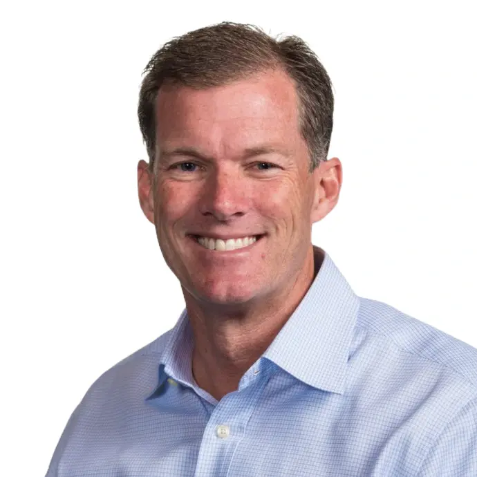 A man with short brown hair, wearing a light blue collared shirt, smiles at the camera against a plain white background.