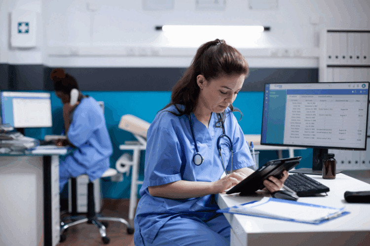 Two healthcare professionals in scrubs work in a medical office; one uses a tablet at a desk, while the other talks on the phone in the background.