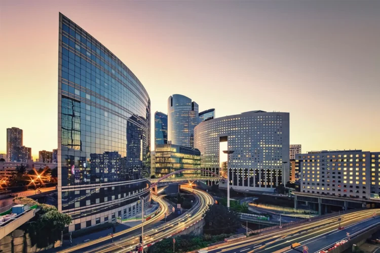 Modern cityscape at sunset showing glass office buildings, busy highways with light trails from cars, and clear sky in the background.