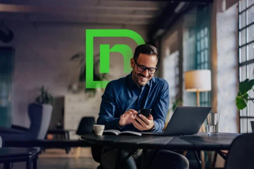A man in glasses sits at a table using a smartphone, with a laptop and notebook in front of him. A large green logo with the letter “n” is overlaid in the background.