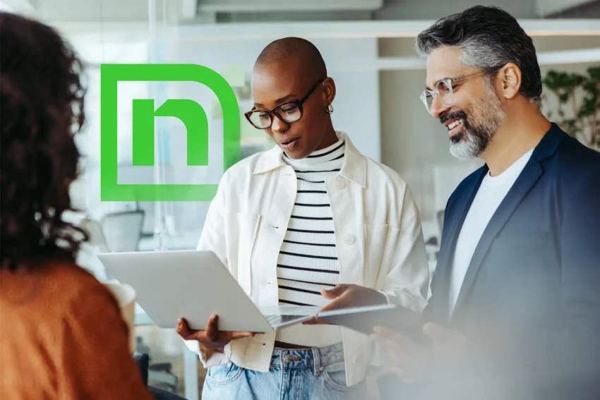 Three people in a modern office setting discuss work; one holds a laptop, another a tablet, with a green logo featuring an "n" visible on the glass wall behind them.