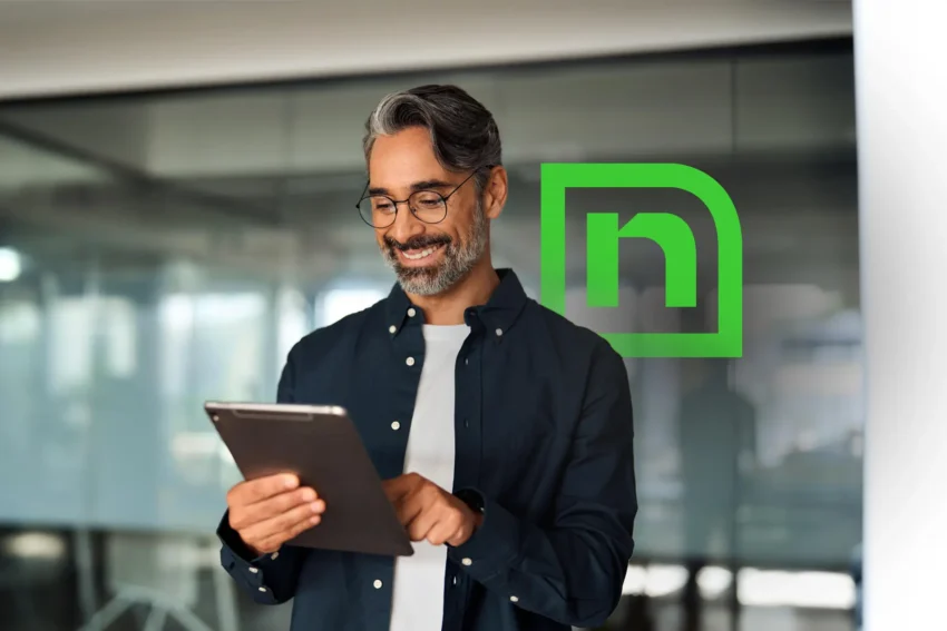 A man with gray hair and glasses uses a tablet while standing indoors. A green tech company logo is visible on the glass wall behind him.
