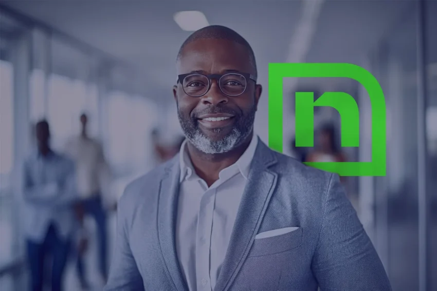 A man in a gray suit and glasses stands indoors, smiling, with a green logo featuring the letter "n" in the background.