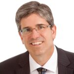 Man with short gray hair and glasses wearing a dark pinstripe suit, white shirt, and patterned tie, smiling at the camera against a white background.