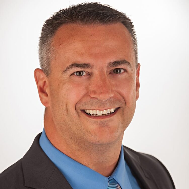 Man in a suit and blue shirt smiling at the camera against a plain white background.