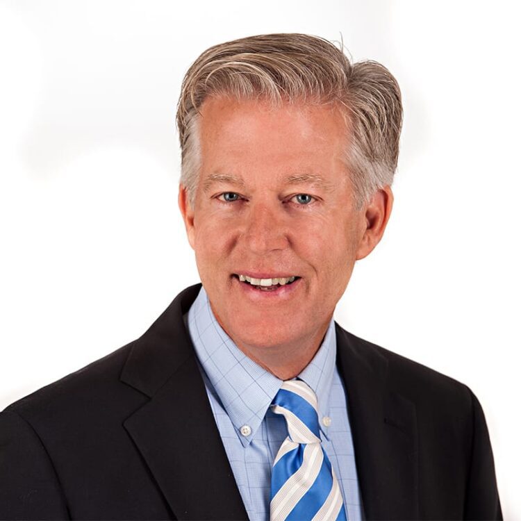 A middle-aged man in a dark suit, light blue shirt, and blue striped tie is smiling at the camera against a white background.