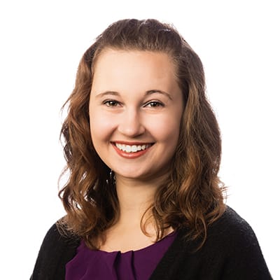 A woman with wavy brown hair, wearing a dark sweater over a purple top, smiles at the camera against a white background.