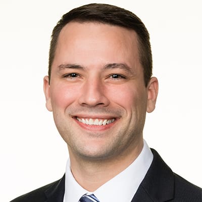 A man with short brown hair wearing a dark suit, white shirt, and striped tie, smiling at the camera against a plain light background.