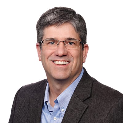 Man with short gray hair and glasses wearing a brown blazer and light blue shirt, smiling at the camera against a white background.