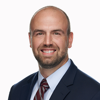 A man with a trimmed beard and short hair wearing a navy suit, light blue shirt, and striped tie, smiling against a plain white background.