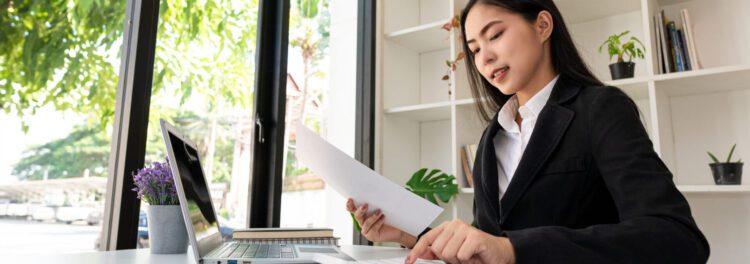 A woman in a black blazer works at a desk with papers, a laptop, and a potted plant in a bright office with large windows, reviewing cloud best practices.