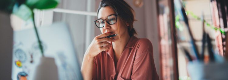 A person wearing glasses and a pink shirt sits at a desk, looking thoughtfully at a laptop while holding a pen to their lips, perhaps researching how to fight inflation.
