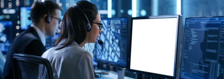 Two people wearing headsets work at computer stations with multiple monitors in a control room, illustrating managed services uses in 2024; one screen is blank.
