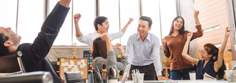 Group of colleagues in an office celebrate together around a table, some raising their fists and smiling enthusiastically, inspired by recent successes with managed services examples.