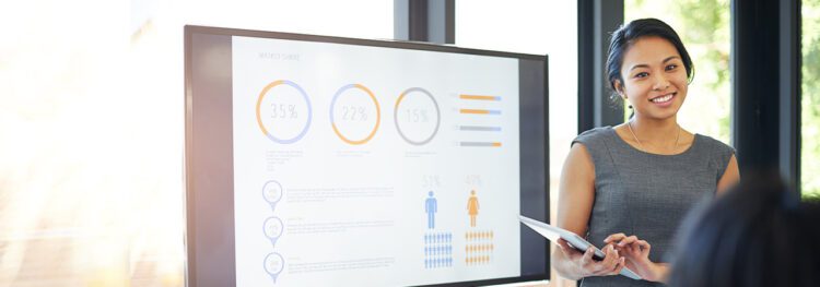 A woman stands next to a large screen displaying charts and graphs, holding a tablet and presenting hr digital transformation data to an audience in a modern office setting.
