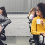 Two people sit at desks in a modern office, discussing intelligent automation examples as they smile at each other. One holds a coffee mug while other colleagues work in the background.