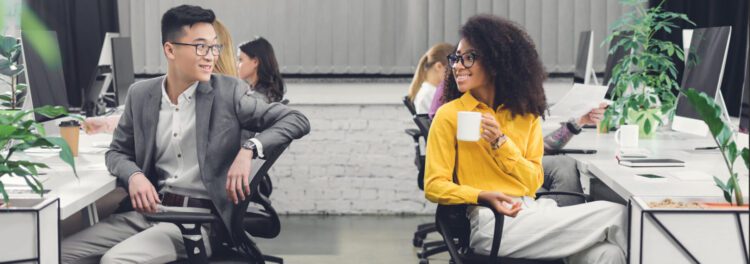 Two people sit at desks in a modern office, discussing intelligent automation examples as they smile at each other. One holds a coffee mug while other colleagues work in the background.