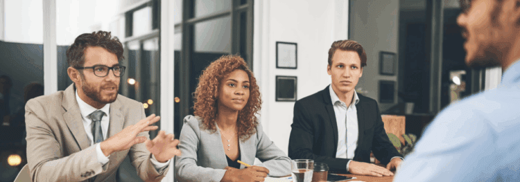 Three people in business attire sit at a table, listening to and interviewing another person whose back is to the camera, discussing HR change management in a modern office setting.
