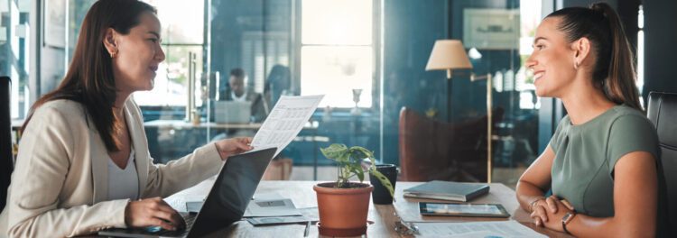 Two women sit across a desk in an office, discussing documents and HR strategy with laptops, papers, and a potted plant on the table.