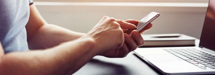 A person focused on digital accessibility uses a smartphone at a desk, with an open laptop and a closed notebook in the background.