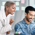 Two people in an office celebrate while looking at a laptop screen, smiling and making fist pump gestures—showing how to be an agent of change. Colorful sticky notes are visible in the background.
