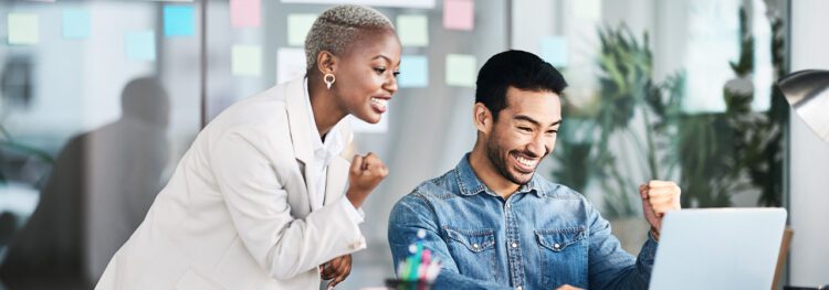 Two people in an office celebrate while looking at a laptop screen, smiling and making fist pump gestures—showing how to be an agent of change. Colorful sticky notes are visible in the background.