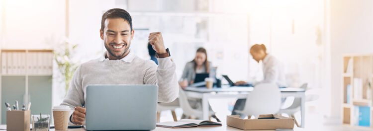 A man sits at a desk with a laptop, smiling and raising his fist in celebration—perhaps enjoying the advantages of cloud storage. Other people work in the background in a bright, modern office.