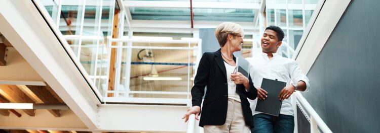 Two colleagues walk down a staircase in a modern office, talking and smiling about hr automation use cases; one holds a clipboard, both appear engaged in conversation.
