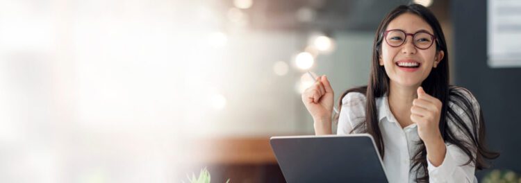 A woman wearing glasses and a white shirt smiles and raises her fists in excitement while sitting at a desk with a tablet, delighted by the possibilities of automating human resources.
