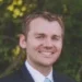 A man in a suit and tie is smiling outdoors with greenery in the background.