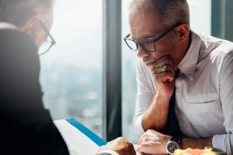 Two men in business attire sit at a table reviewing documents, with one man resting his chin on his hand and wearing glasses.