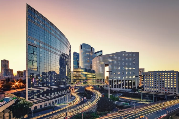 Modern glass office buildings with curved facades at sunset, with light trails from cars on roads and overpasses in the foreground.