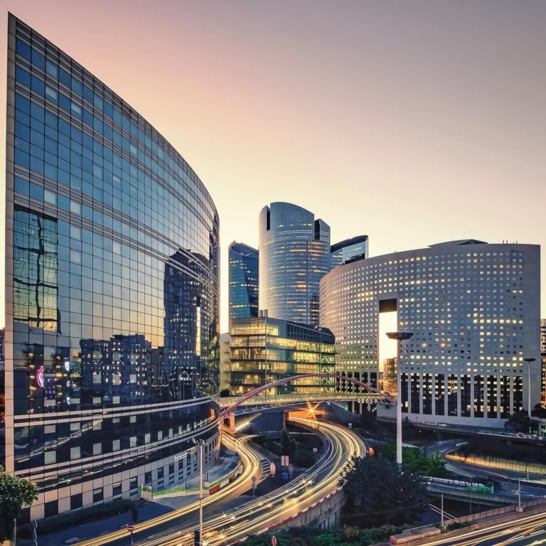 Modern cityscape with tall glass office buildings at sunset, showing reflections on the facades and light trails from vehicles on curving roads below.
