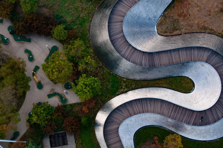 Aerial view of a winding, elevated wooden walkway curving through a park with trees and a small mini-golf course below.