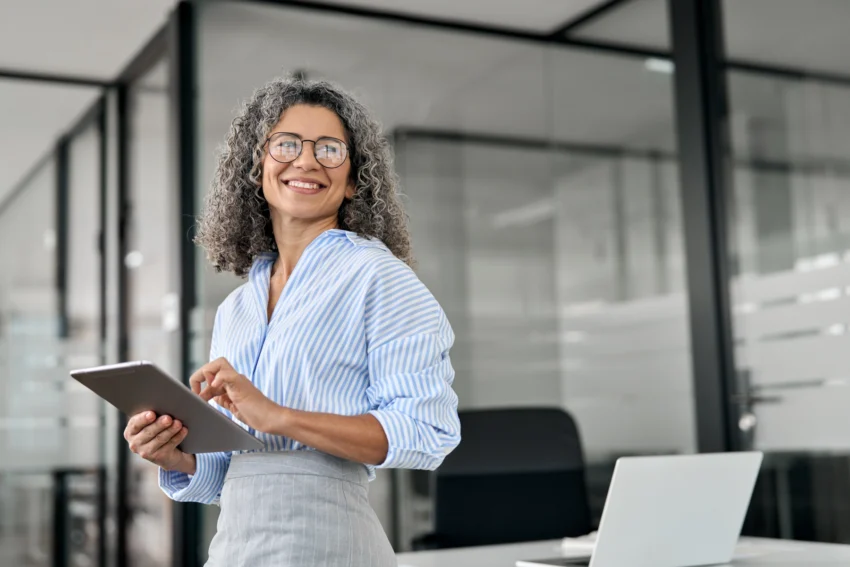 Smiling woman with curly gray hair in business attire stands in an office holding a tablet, with a laptop on the desk in the background.