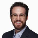 Man with short brown hair, beard, and glasses, wearing a dark blazer and light-colored checkered shirt, smiling at the camera against a plain white background.