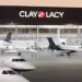 A row of private jets is parked on the tarmac in front of a building with the sign "Clay Lacy.