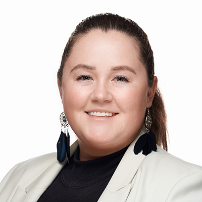 A woman with brown hair in a ponytail, wearing a white blazer, black top, and large feather earrings, smiles at the camera against a plain white background.