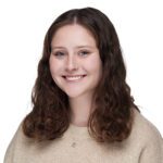 A young woman with wavy brown hair and a beige sweater smiles at the camera against a white background.