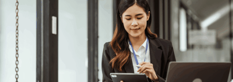 A woman in business attire uses a digital tablet and stylus while sitting at a desk with a laptop, embracing 2025 HR trends in a modern office setting.