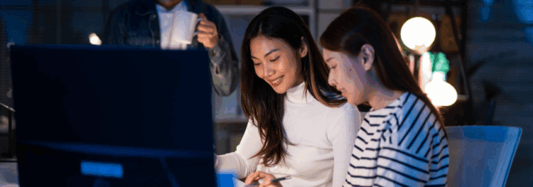 Two women sit together at a desk looking at documents powered by IDP AI in front of a computer screen, while a person in the background holds a mug.