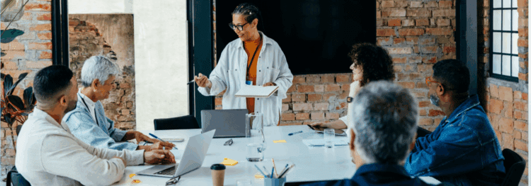 A group of six people sit around a table in a modern office while one person stands and presents with a clipboard, discussing ocr vs idp. Laptops and papers are on the table.