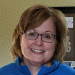 A woman with short light brown hair and glasses smiles at the camera, wearing a blue top. A framed item is visible in the background.