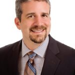 A man with short gray hair and a beard, wearing a dark suit jacket, checkered shirt, and striped tie, smiles at the camera against a plain white background.