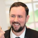 A man with short dark hair and a beard, wearing a suit and tie, smiles indoors with blurred windows in the background.