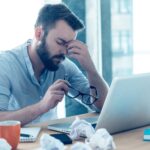 Man sitting at a desk with a laptop, holding his glasses and rubbing his eyes, surrounded by crumpled papers and office supplies—a clear sign of the challenges faced during Change Management.