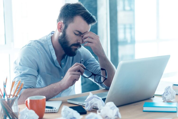 Man sitting at a desk with a laptop, holding his glasses and rubbing his eyes, surrounded by crumpled papers and office supplies—a clear sign of the challenges faced during Change Management.